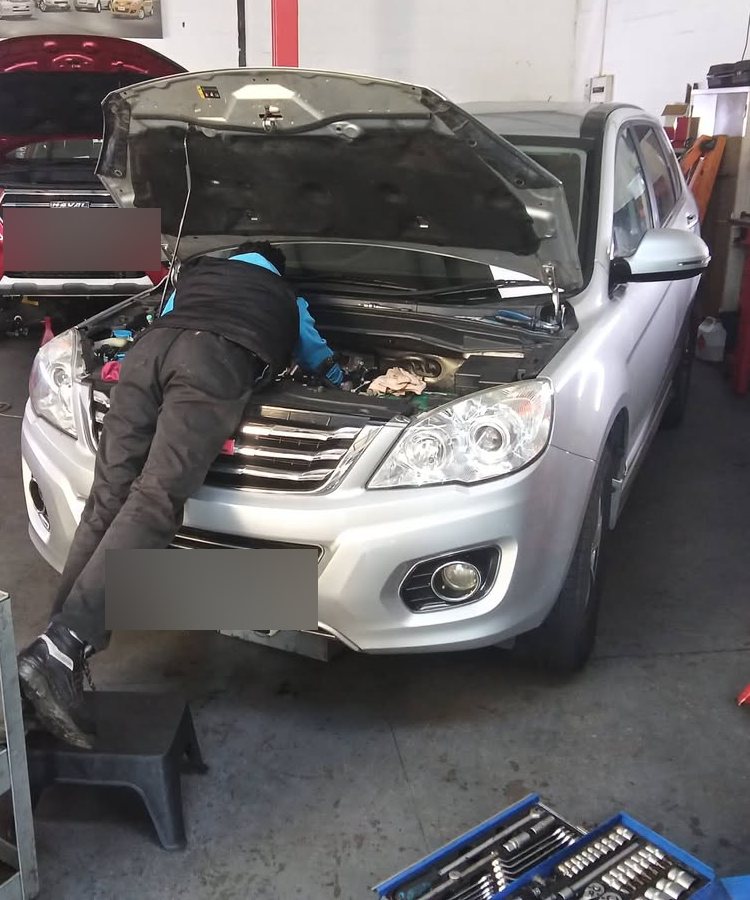An H2 technician working under the bonnet of a silver GWM in the Brackenfell workshop, with a red Haval on the lift behind it.